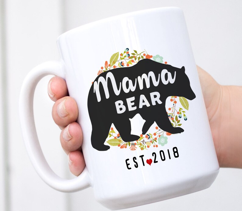 May include: White ceramic mug featuring a black silhouette of a bear and the text "Mama Bear Est. 2018" encircled by a floral wreath.
