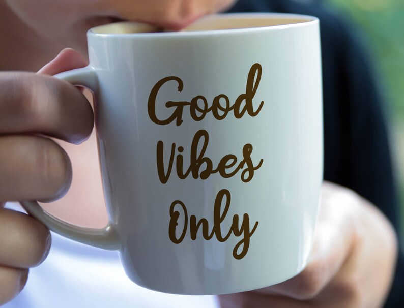 May include: A white ceramic mug with the words "Good Vibes Only" in a brown script. The mug has a handle and is being held by a person. The mug is filled with a light-colored liquid.