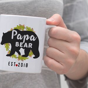 May include: White ceramic mug featuring a black silhouette of a bear and the text "Papa Bear Est. 2018" encircled by a green wreath.