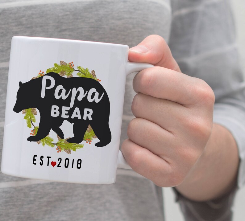 May include: White ceramic mug featuring a black silhouette of a bear and the text "Papa Bear Est. 2018" encircled by a green wreath.