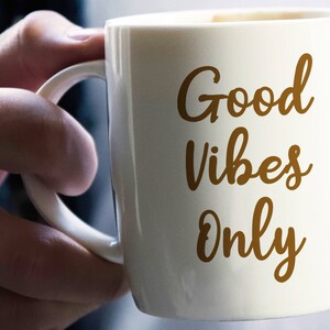 May include: A white ceramic mug with the words "Good Vibes Only" in a brown script. The mug has a curved handle and is held by a person's hand. The mug is filled with a dark liquid, possibly coffee or tea.