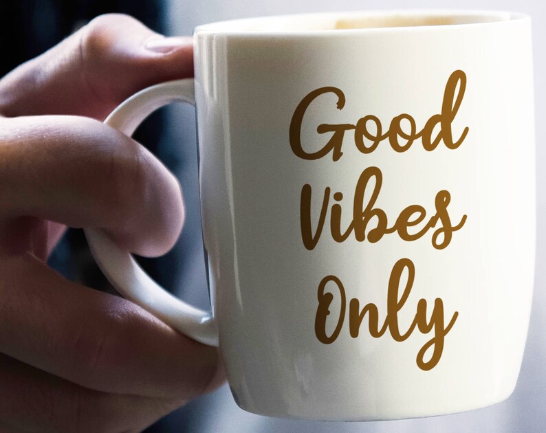 May include: A white ceramic mug with the words "Good Vibes Only" in a brown script. The mug has a curved handle and is held by a person's hand. The mug is filled with a dark liquid, possibly coffee or tea.