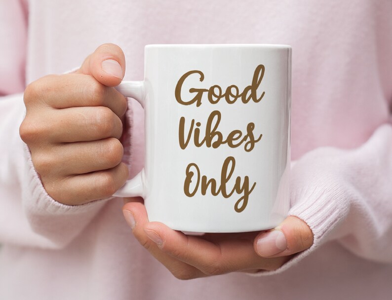 May include: A white ceramic mug with the words "Good Vibes Only" in a brown script font. The mug is held by a person wearing a pink sweater. The mug has a large handle and is likely used for coffee or tea.