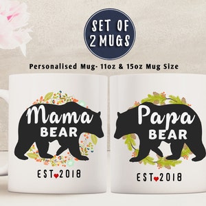 May include: Set of two white ceramic mugs featuring black bear silhouettes and floral wreaths. The mugs are personalised with "Mama Bear" and "Papa Bear" and the year "EST 2018".
