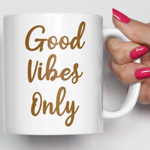 May include: White ceramic mug with the words "Good Vibes Only" in a brown script font. The mug has a curved handle and is held by a person with pink nail polish.