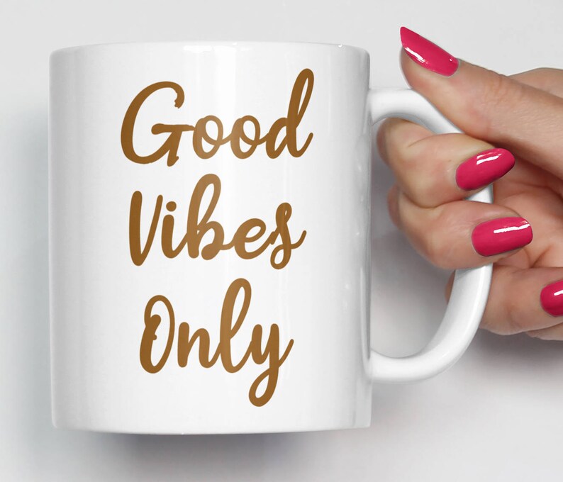 May include: White ceramic mug with the words "Good Vibes Only" in a brown script font. The mug has a curved handle and is held by a person with pink nail polish.