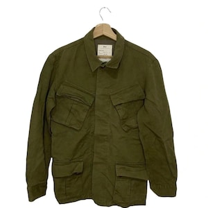 May include: Olive green jacket with four front pockets and a collared neckline. The jacket is hanging on a wooden hanger. The fabric appears to be a durable, possibly cotton, material. The jacket has a button-up front.