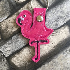 Könnte beinhalten: Rosafarbener Flamingokettenanhänger mit schwarzen Nähten und einem silbernen Metallring. Der Flamingo steht auf einem Bein und hat den Kopf zur Seite gedreht.