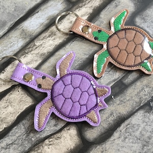 May include: Two felt turtle keychains. One is purple with a brown head and feet. The other is brown with a green head and feet.