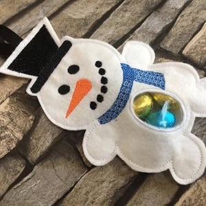 May include: White felt snowman ornament with a black top hat, blue scarf, and a clear plastic window showing colorful candy.