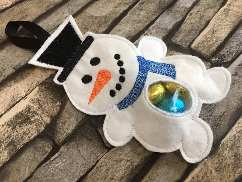 May include: White felt snowman ornament with a black top hat, blue scarf, and a clear plastic window showing colorful candy.