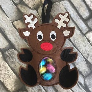 May include: A brown felt reindeer ornament with a clear plastic window showing colorful chocolate eggs. The reindeer has a red nose and black eyes.