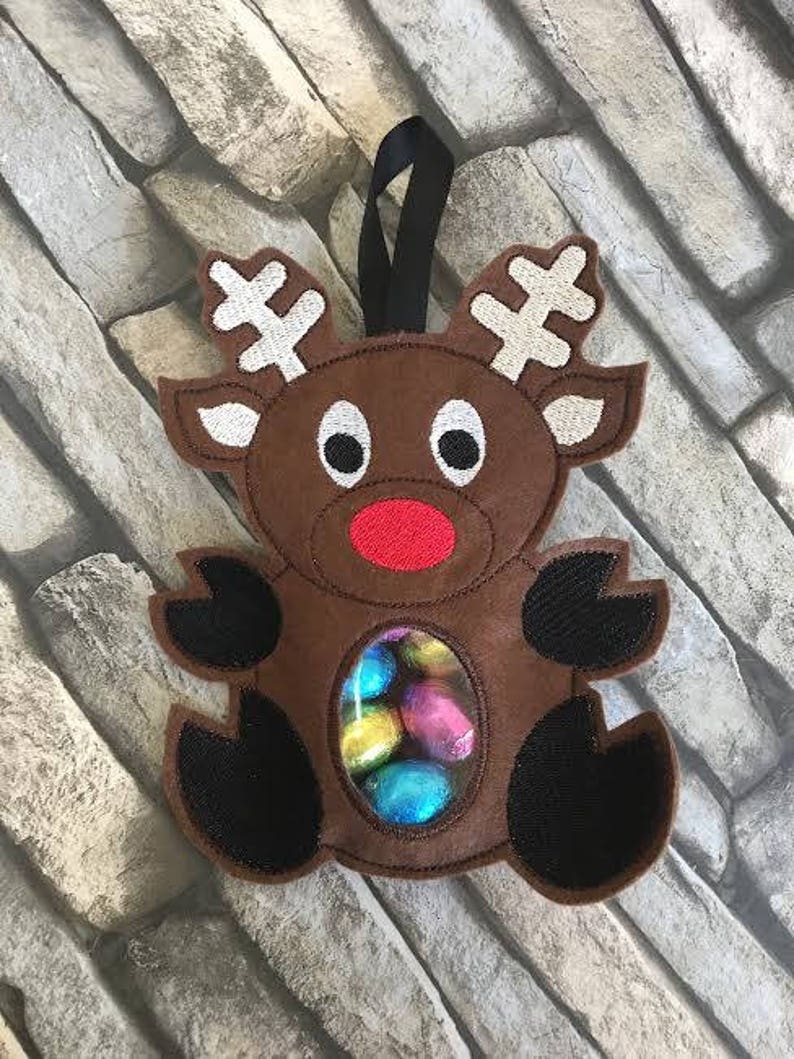 May include: A brown felt reindeer ornament with a clear plastic window showing colorful chocolate eggs. The reindeer has a red nose and black eyes.