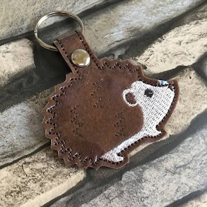 Peut inclure: Un porte-clés en cuir marron avec un motif de hérisson brodé. Le hérisson a un visage blanc et un corps marron avec des coutures en zigzag. Le porte-clés comprend un anneau métallique et une lanière en cuir avec un bouton argenté.