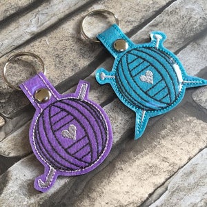 May include: Two yarn ball keychains, one purple and one blue, with silver knitting needles and a silver heart on each.