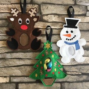 May include: Three felt Christmas ornaments, a reindeer, a snowman, and a Christmas tree, each with a pocket for treats. The reindeer is brown with a red nose, the snowman is white with a black hat and blue scarf, and the Christmas tree is green with red ornaments and a brown base.