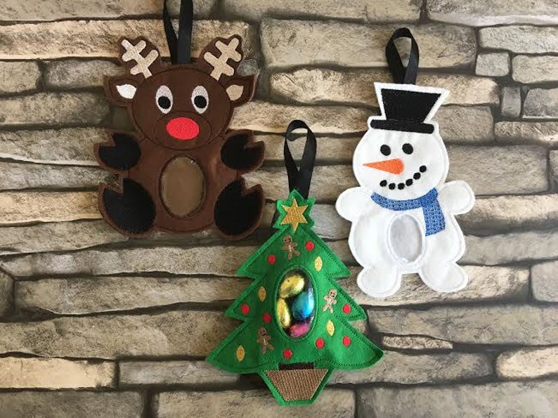 May include: Three felt Christmas ornaments, a reindeer, a snowman, and a Christmas tree, each with a pocket for treats. The reindeer is brown with a red nose, the snowman is white with a black hat and blue scarf, and the Christmas tree is green with red ornaments and a brown base.