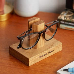 Może przedstawiać: Drewniany uchwyt na okulary z wygrawerowanym napisem "Grandpa's Glasses" z przodu. Uchwyt ma dwa pionowe podpory do trzymania okular&oacute;w, a para okular&oacute;w w czarnych oprawkach spoczywa w uchwycie.