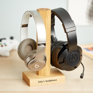 Könnte beinhalten: Ein hölzerner Kopfhörerständer mit zwei Kopfhörerpaaren. Ein Paar ist silber und weiß, das andere ist schwarz. Der Ständer ist mit dem Text "Toby's headphones" graviert.