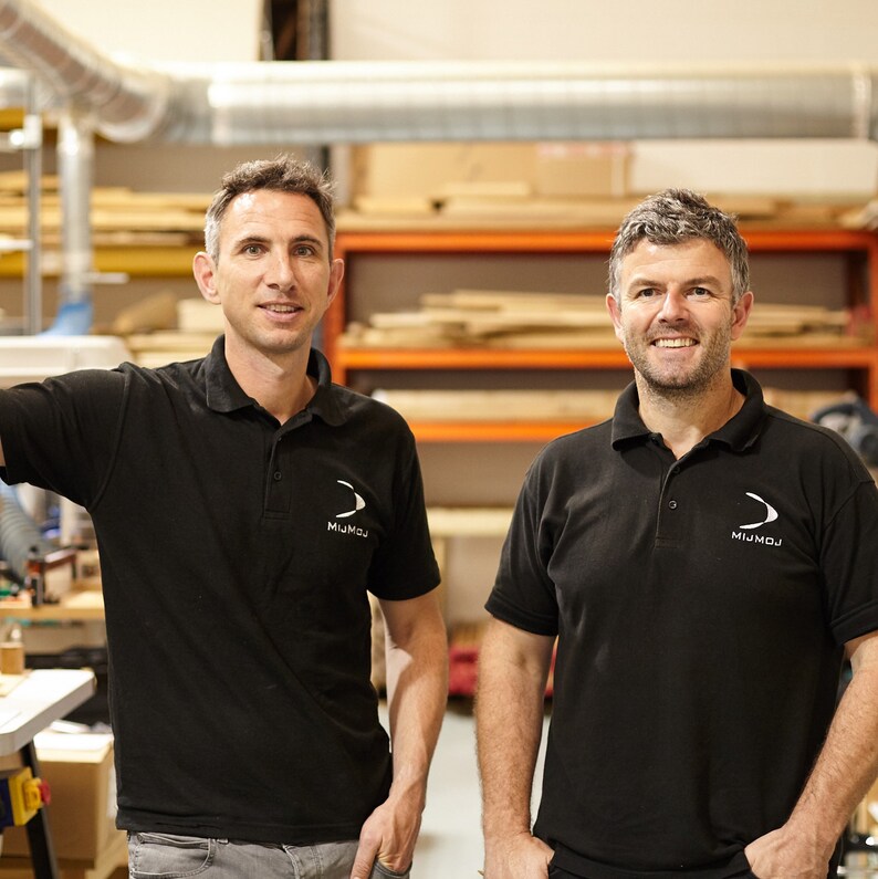 May include: Two men wearing black polo shirts with the "MIJMOJ" logo, standing in a workshop. The background includes wooden shelves and industrial equipment. The men are smiling at the camera.