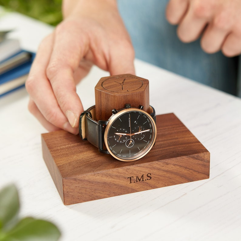 May include: A black-faced watch with a gold-coloured bezel and black leather strap sits on a wooden watch stand. The stand is engraved with the initials "T.M.S." The watch stand is made of dark wood.