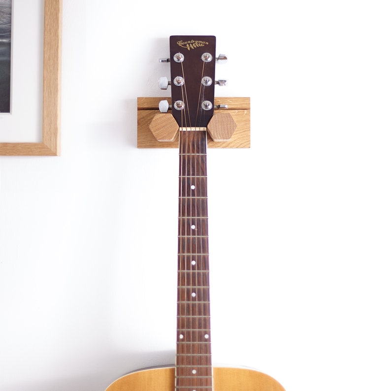 May include: A light-coloured wooden guitar wall mount featuring a rectangular backplate and two hexagonal supports holding the dark brown headstock of an acoustic guitar with the brand name Countryman printed in gold lettering.
