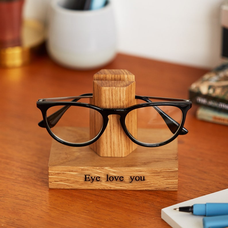 Może przedstawiać: Drewniany stojak na okulary z wygrawerowanym tekstem "Eye love you" na przodzie. Stojak wykonany jest z naturalnego drewna i ma prosty, elegancki design.