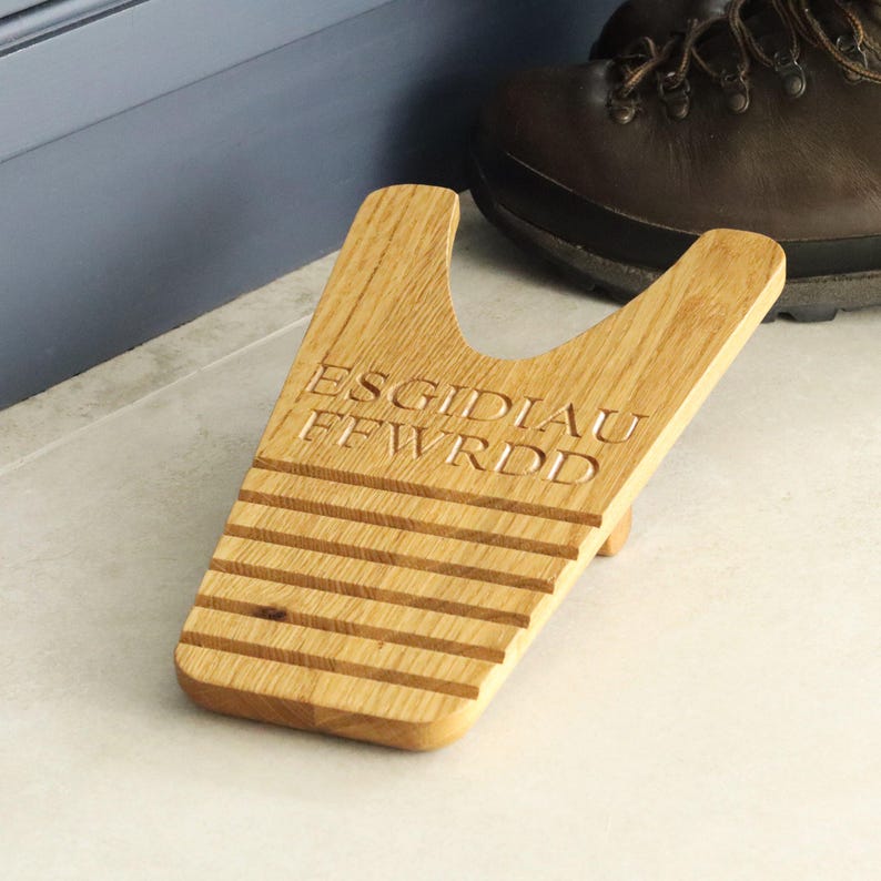 Pu&ograve; includere: Un cavatacchi in legno con il testo inciso "ESGIDIAU FEWRDD". Il cavatacchi &egrave; realizzato in legno chiaro e ha una parte superiore a forma di Y con scanalature orizzontali. Uno stivale marrone &egrave; sullo sfondo.