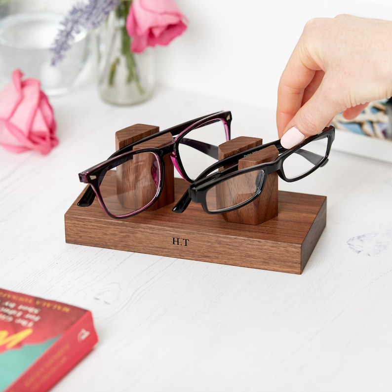 May include: A wooden glasses holder with three slots, holding two pairs of glasses. One pair has black frames with pink accents, and the other has black frames. The holder is engraved with the initials "H.T."