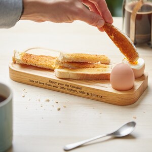 May include: A wooden breakfast serving tray with a personalized message: "Enjoy your dip Grandad, lots of love from your little soldiers, Alfie & Billie xx". The tray has a small indentation for a boiled egg and a larger area for toast.