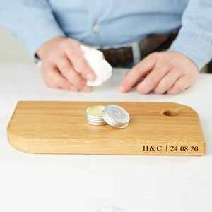 May include: A wooden cutting board with a rounded edge and a small hole. Two small, open silver tins with a yellow substance inside sit on the board. The board has the engraved text "H&C | 24.08.20".