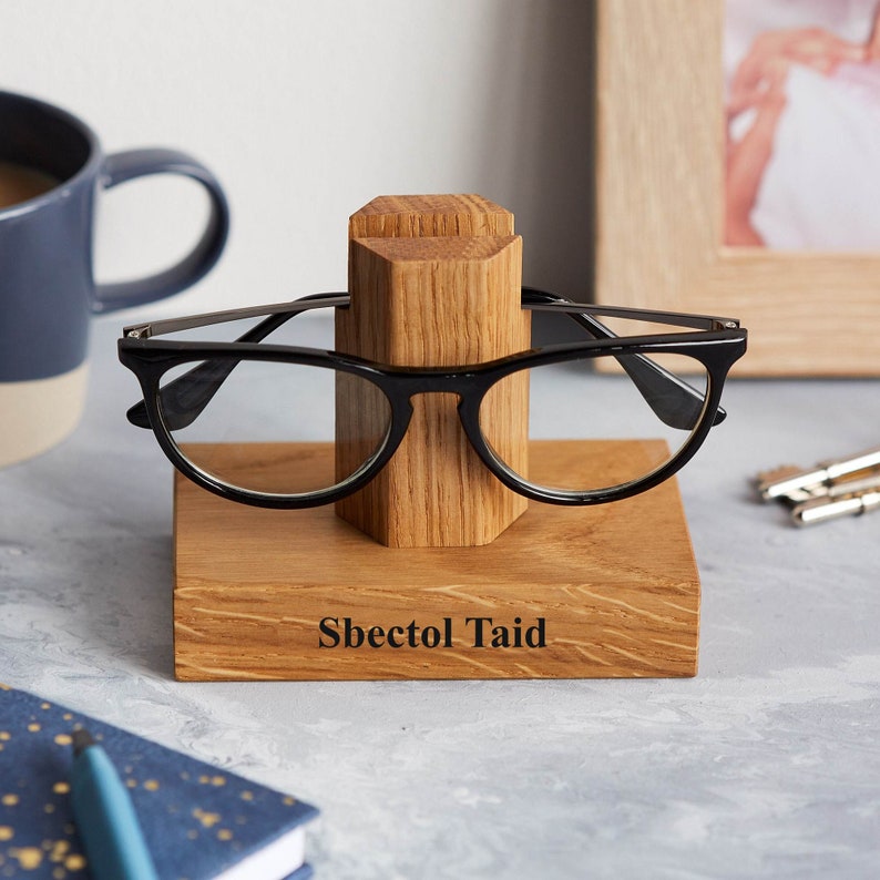 Może przedstawiać: Drewniany stojak na okulary z personalizowanym napisem "Sbectol Taid". Stojak wykonany jest z jasnobrązowego drewna i ma prosty design. Czarne okulary leżą na stojaku.