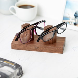 May include: A wooden glasses holder with two pairs of glasses resting on it. The holder is engraved with the initials "H.T."