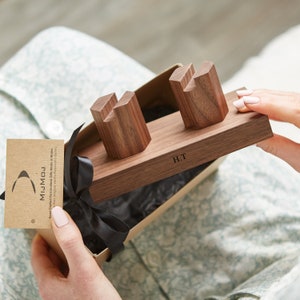 May include: A wooden phone stand with two slots for phones. The stand is made of walnut wood and has a personalized inscription that reads "H.T.". The stand is in a gift box with a black ribbon.