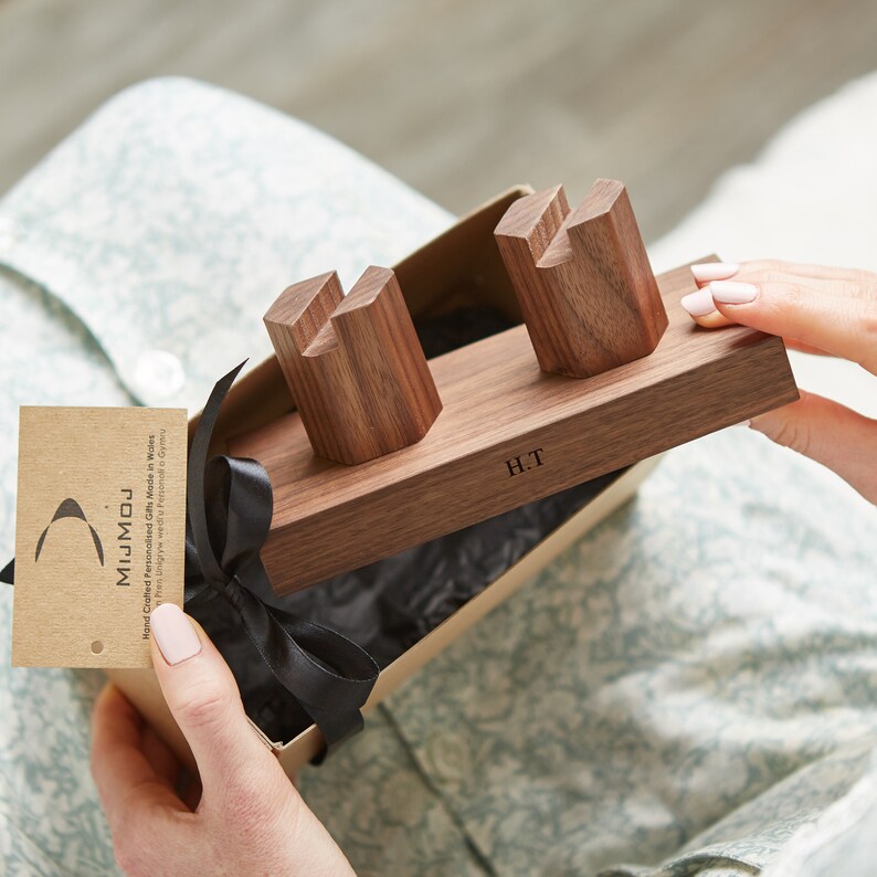 May include: A wooden phone stand with two slots for phones. The stand is made of walnut wood and has a personalized inscription that reads "H.T.". The stand is in a gift box with a black ribbon.