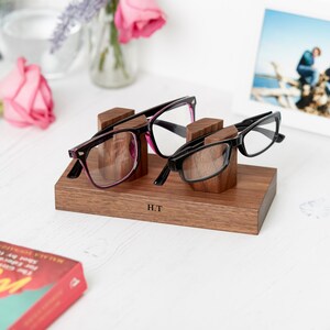May include: A wooden eyeglasses holder with two pairs of glasses. One pair has a black frame with pink accents, and the other has a black frame. The holder is engraved with the initials "H.T."