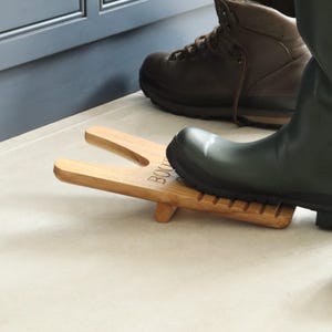 Pu&ograve; includere: Un cavastivali in legno con una scanalatura per il tallone e un'estremit&agrave; a forcella per tenere lo stivale. Uno stivale di gomma verde poggia sul cavastivali, con uno scarponcino da trekking in pelle marrone sullo sfondo. La parola "BOOT" &egrave; visibile.