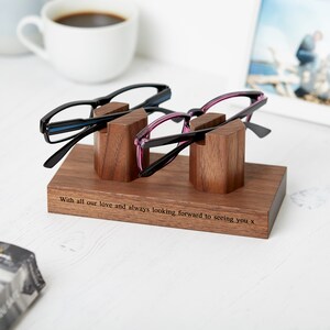 May include: A rectangular wooden glasses stand with two upright supports holding a pair of black eyeglasses and a pair of purple eyeglasses. The base features the engraved text: With all our love and always looking forward to seeing you x.