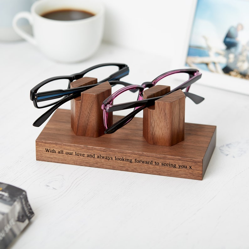 May include: A rectangular wooden glasses stand with two upright supports holding a pair of black eyeglasses and a pair of purple eyeglasses. The base features the engraved text: With all our love and always looking forward to seeing you x.