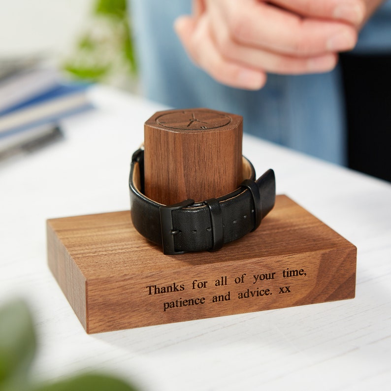 May include: A wooden watch stand with a black leather watch. The stand has a rectangular base with engraved text and a hexagonal top to hold the watch. The watch has a black leather strap and a black buckle. The text on the base reads, "Thanks for all of your time, patience and advice. xx."
