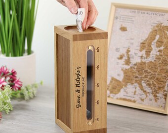 Op de afbeelding: Een houten spaarpot voor een bruiloft met een transparant venster dat het spaarniveau aangeeft. De doos heeft een gleuf aan de bovenkant om geld in te doen. De tekst "Simon & Natasha's WEDDING FUND" staat erop. Een hand steekt een biljet erin.