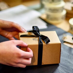 May include: A small, brown cardboard gift box tied with a black satin ribbon. The box has the text "MIJ Hand Crafted and Design" printed on the top. The box is being held by a person.