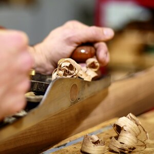 May include: A hand plane in action, creating wood shavings. The plane is held by a hand, with the blade cutting into the wood. The shavings are light brown and curled. The image shows a woodworking process.
