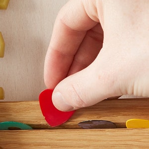 May include: A hand placing a red guitar pick into a wooden guitar pick holder. The holder has slots for multiple picks and is filled with picks of various colours.