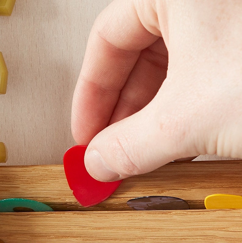 May include: A hand placing a red guitar pick into a wooden guitar pick holder. The holder has slots for multiple picks and is filled with picks of various colours.