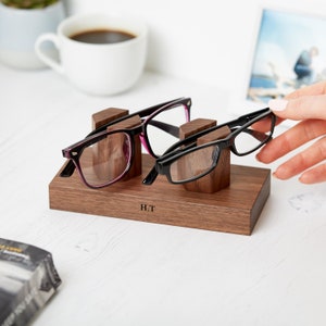 May include: A rectangular dark wood glasses stand featuring two upright supports for holding eyewear. The base is engraved with the letters H.T. Two pairs of glasses are resting on the stand, one with black and pink frames and one with black frames.