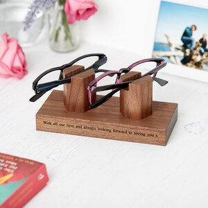 May include: A wooden eyeglasses holder with two pairs of glasses. The holder is rectangular with two vertical blocks to hold the glasses. The glasses have black frames with blue and pink accents. The holder has the text "With all our love and always looking forward to seeing you x."