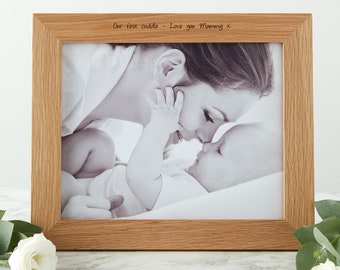 May include: A rectangular wooden picture frame with a black and white photo of a mother and baby. The frame has the inscription "Our first cuddle - Love you Mummy x". The frame is made of light brown wood.