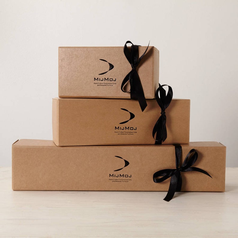 Op de afbeelding: Drie gestapelde rechthoekige kartonnen dozen, vastgebonden met zwarte satijnen linten. Elke doos heeft een zwart logo en de tekst "MIJMOJ Hand Crafted Personalized Gifts and Bespoke Furniture."