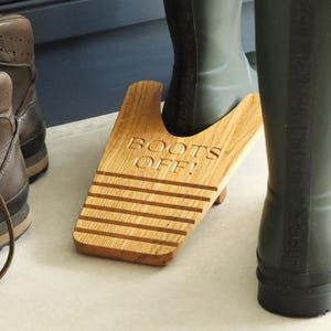 May include: A wooden boot jack with the words "BOOTS OFF!" engraved on the surface. The boot jack is made of light-colored wood and has horizontal grooves. Green rubber boots are in use with the boot jack.